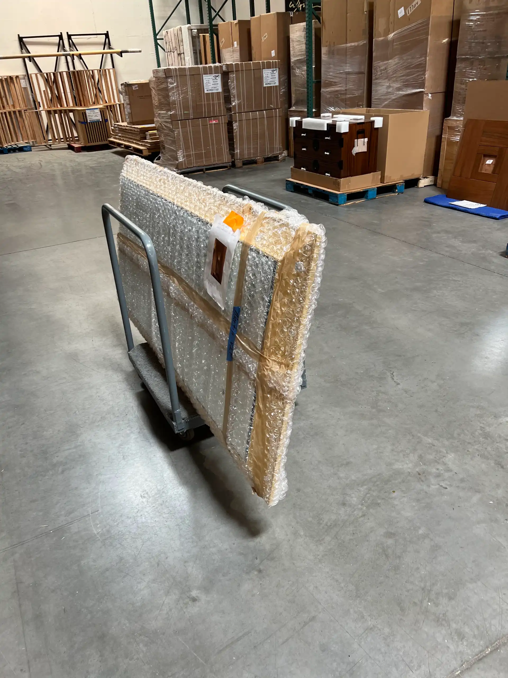 A bubble-wrapped panel tagged and moved by cart past racked, palletized inventory