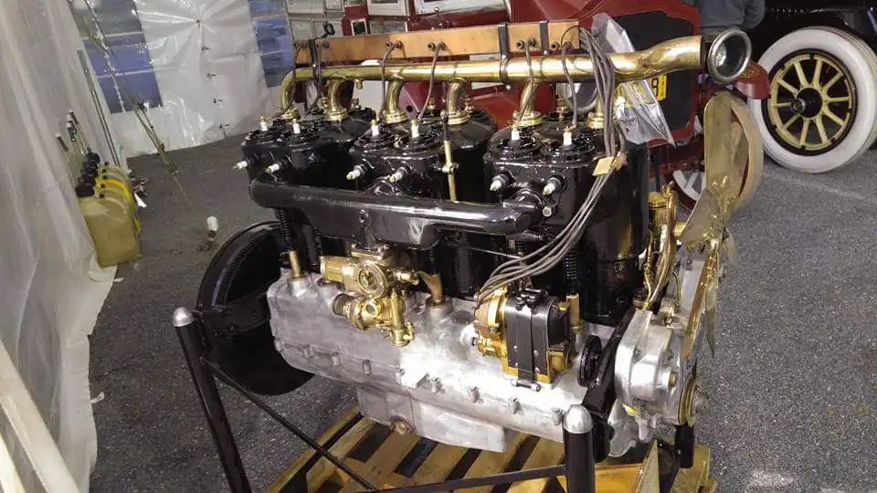 A restored brass-era automobile engine on a rolling stand beside its vehicle